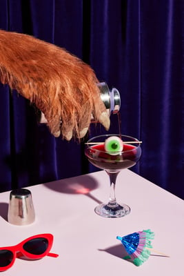 Crop of werewolf hairy hand spilling red beverage into cocktail glass with eye on pink table with purple velvet curtain background