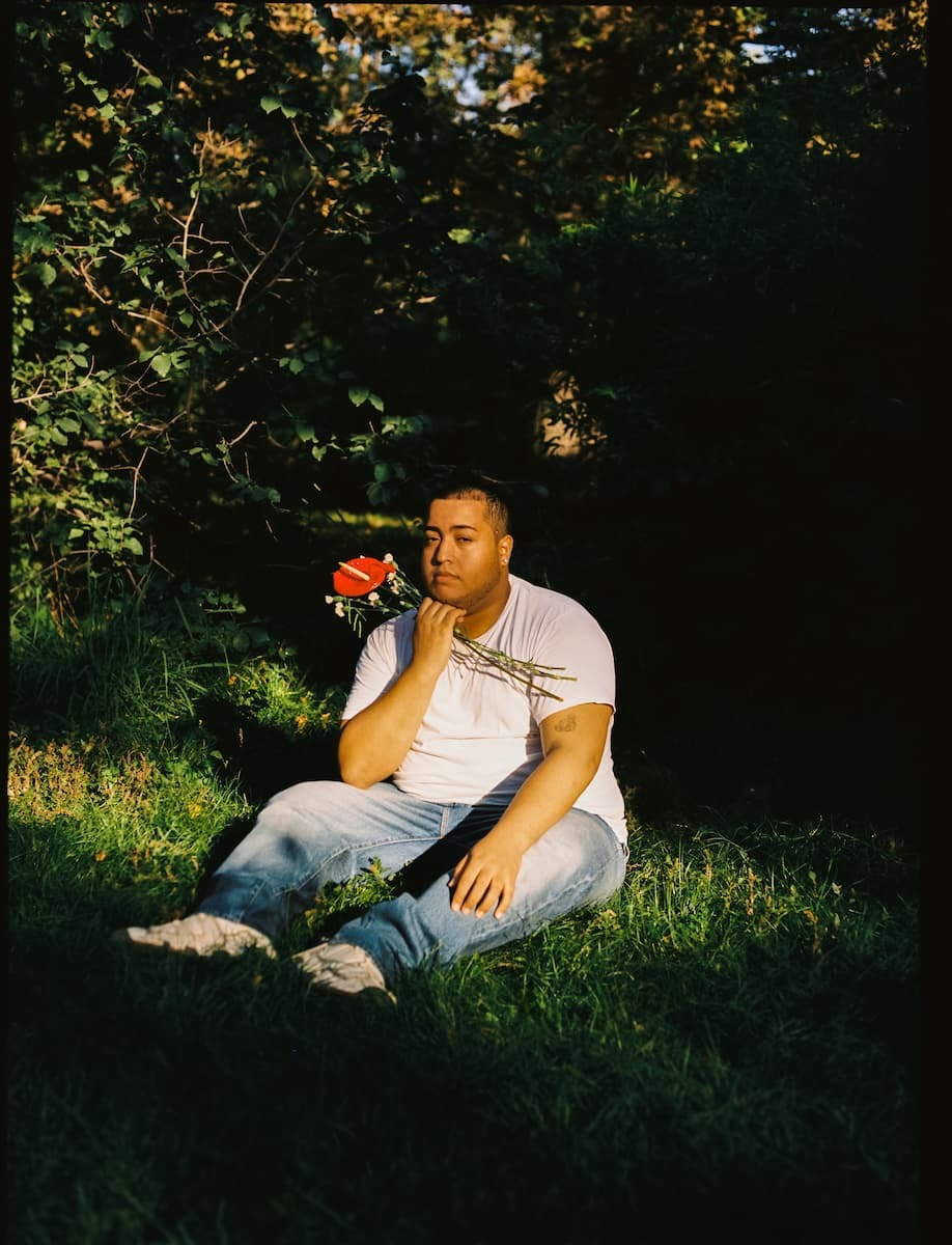 Black Plus-Size Guy Sitting On The Grass Looking Into The Camera. Portrait of plus size black boy sitting on the grass of a park in warm afternoon light looking at camera, wearing basic white t-shirt and jeans. Holding red and white-flowers in his hand. Shot on medium format Kodak Portra film stock.