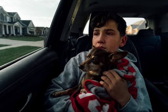 A teen boy is holding the family's dog on a final ride to the vet. The boy has tears streaming down his face while looking out the window.